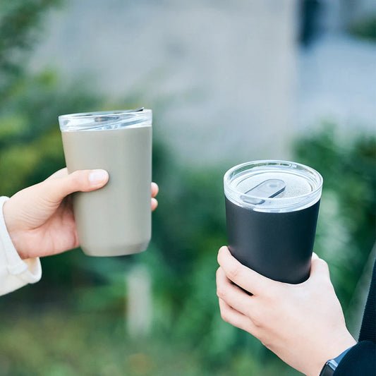 Two hands holding insulated travel mugs outdoors with a blurred green background